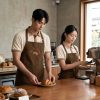 Mẫu đồng phục quán cafe gồm áo thun polo màu be phối tạp dề nâu có in logo cho nhân viên phục vụ và pha chế chuyên nghiệp.